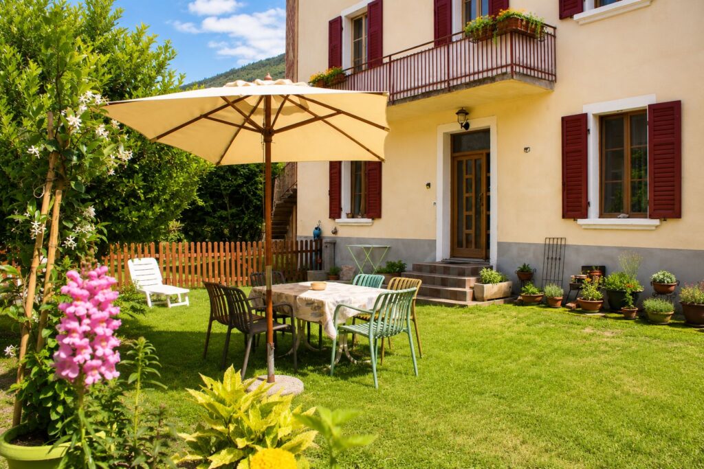 Zona pranzo nel giardino con tavolo, sedie, ombrellone e piante fiorite.