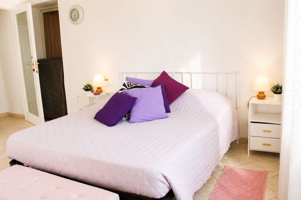 Vista della camera matrimoniale Lilla con letto, cuscini nelle tonalità del viola, comodini bianchi e finestra luminosa.