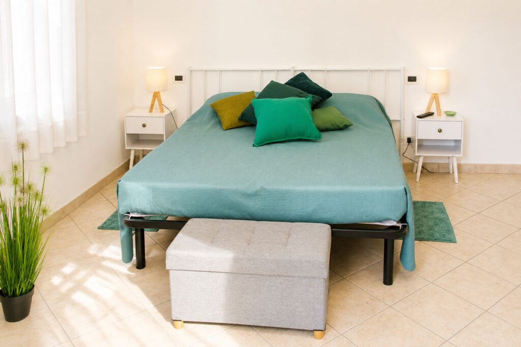 Letto matrimoniale con coperta verde, cuscini colorati e comodini con lampade.